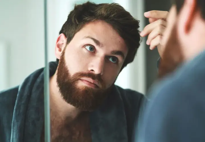 Hombre con barba revisando su pelo graso frente al espejo, con una bata puesta.