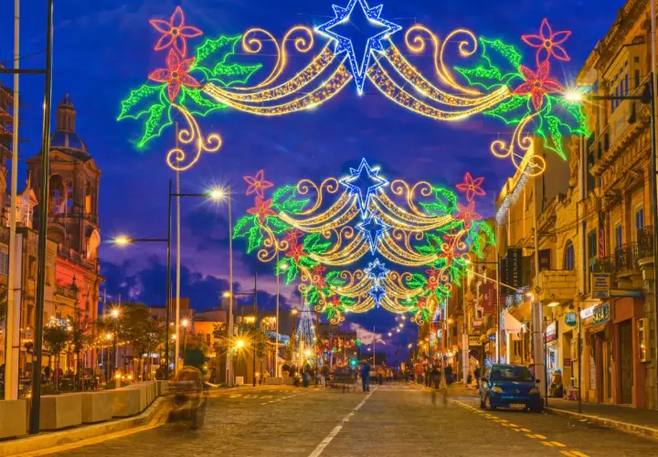 Malta w grudniu rozświetlona świątecznymi iluminacjami. Ulica ozdobiona girlandami z gwiazd i kwiatów, w tle kościół i sklepy.