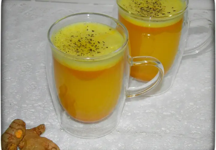 Zwei Gläser mit leuchtend gelbem Kurkuma-Wasser, bestreut mit schwarzem Pfeffer. Daneben liegen frische Kurkumawurzeln. Ein einfaches Rezept für Wohlbefinden.