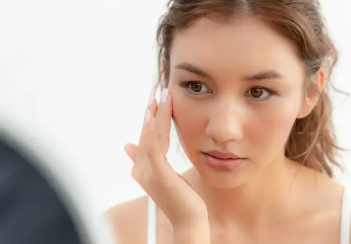 Une jeune femme examine son visage, préoccupée par la sécheresse cutanée. Sa peau semble tendue et manque d'hydratation.