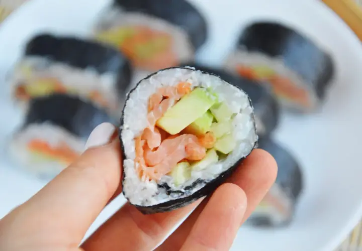 Ręka trzyma sushi z łososiem wędzonym na gorąco, awokado i ogórkiem.