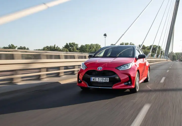 Czerwony Toyota Yaris Hybrid pędzi po moście w słoneczny dzień.
