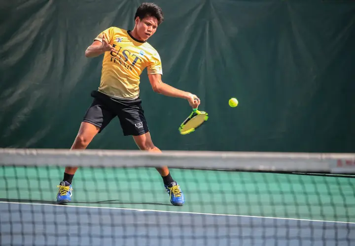 Młody tenisista w żółtej koszulce UST TENNIS uderza piłkę, walcząc o każdy break point.
