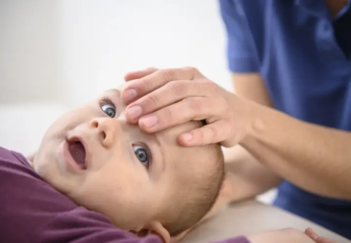 Ein Baby nach Osteopathie, wie ausgewechselt, mit weit geöffnetem Mund und großen Augen, während eine Hand sanft seine Stirn berührt.