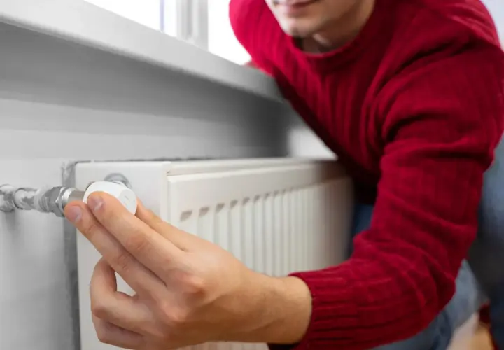 Mężczyzna w czerwonym swetrze reguluje termostat grzejnika, zastanawiając się, czy zakręcać grzejniki na noc, by oszczędzić energię.