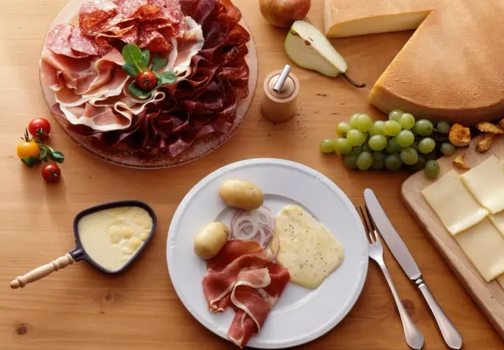 Une table dressée pour une abondance raclette : charcuteries, fromages, pommes de terre, raisins et poêlon de fromage fondu.