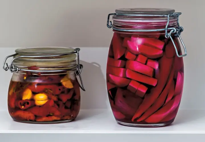 Två glasburkar med inlagd paprika och andra grönsaker. Den större burken innehåller röda, avlånga bitar, troligen inlagd paprika.