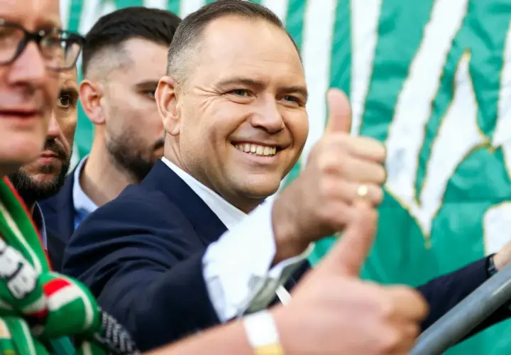 Uśmiechnięty mężczyzna w garniturze, z kciukiem w górze, na tle zielono-białej flagi. Widać kibiców Lechii Gdańsk.