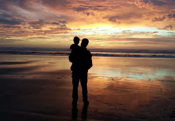 Silhouette di un padre che porta in braccio il figlio sulla spiaggia al tramonto. Un momento di gioia che celebra la festa del papà nel mondo.
