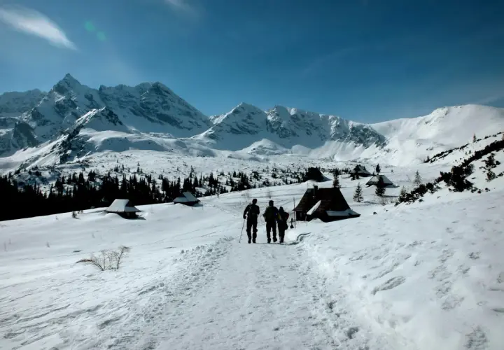 Trzej turyści idą szlakiem przez zaśnieżone Tatry zimą dla średniozaawansowanych, mijając góralskie chatki.