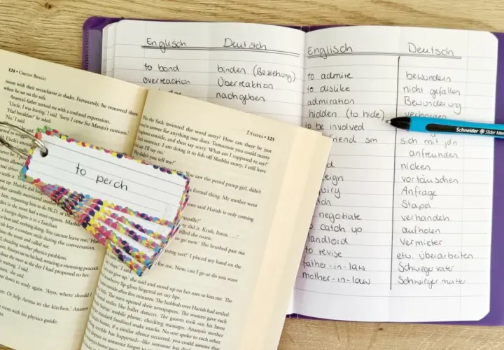 Ein offenes digitales Vokabelheft mit englischen und deutschen Wörtern, daneben ein Buch und Karteikarten zum Lernen.