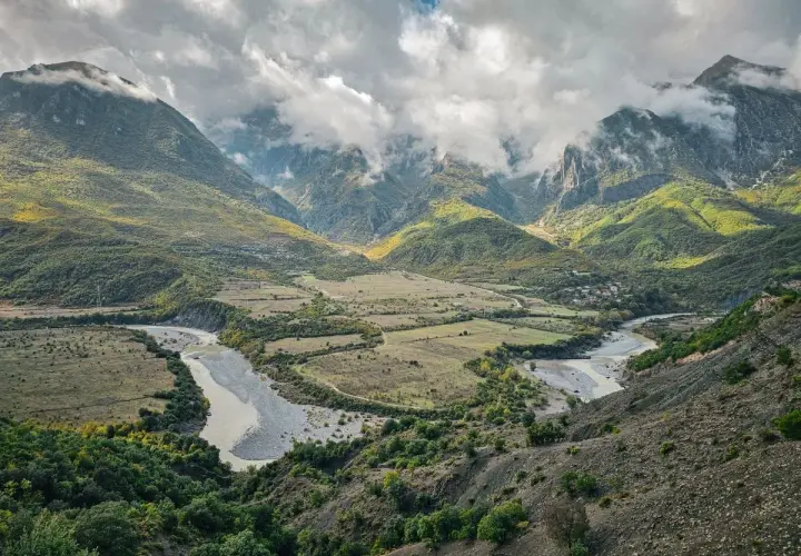 Ein malerischer Fluss schlängelt sich durch ein grünes Tal in Albanien, umgeben von bewaldeten Bergen unter dramatischen Wolken.