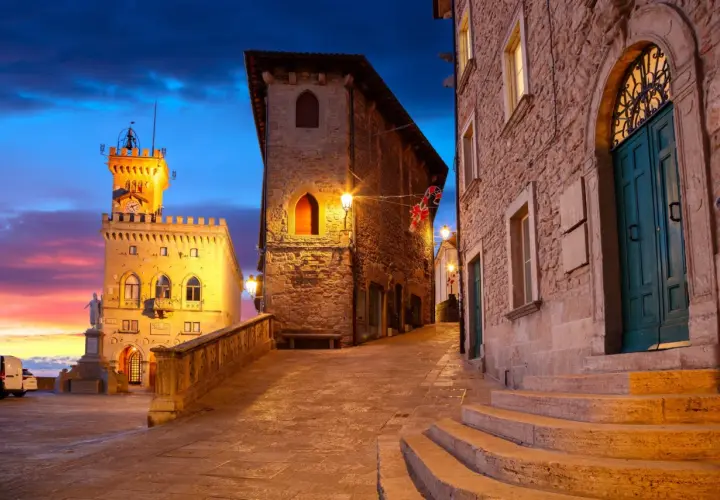 Wieczorna panorama San Marino, z wieżą i kamiennymi budynkami oświetlonymi ciepłym światłem. Niebo mieni się odcieniami różu i błękitu.
