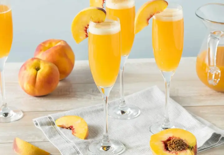 Tre glas med en gyllene drink, garnerade med persikoklyftor. Persikor och en karaff med drinken syns också.