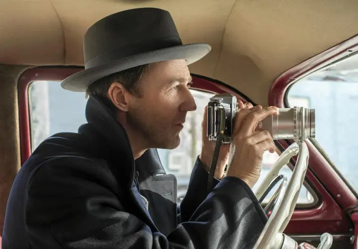 Edward Norton w filmach, w kapeluszu i płaszczu, siedzi za kierownicą samochodu i robi zdjęcie starym aparatem.