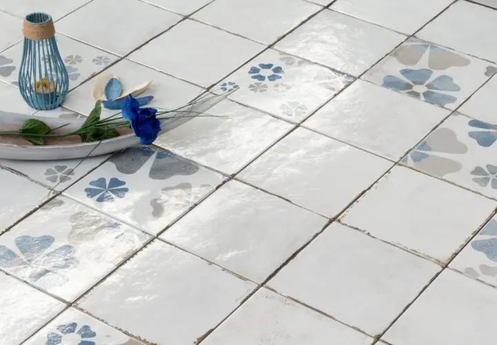 Pavimento con piastrelle quadrate, alcune decorate con fiori blu e grigi, altre bianche. Un vassoio in legno con un fiore blu e un portacandele blu completano la scena, mostrando la differenza tra mattonella e piastrella.