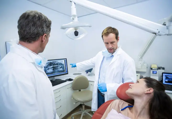 Dwóch dentystów omawia zdjęcie rentgenowskie z pacjentką, zastanawiając się, ile zarabia dentysta NFZ.