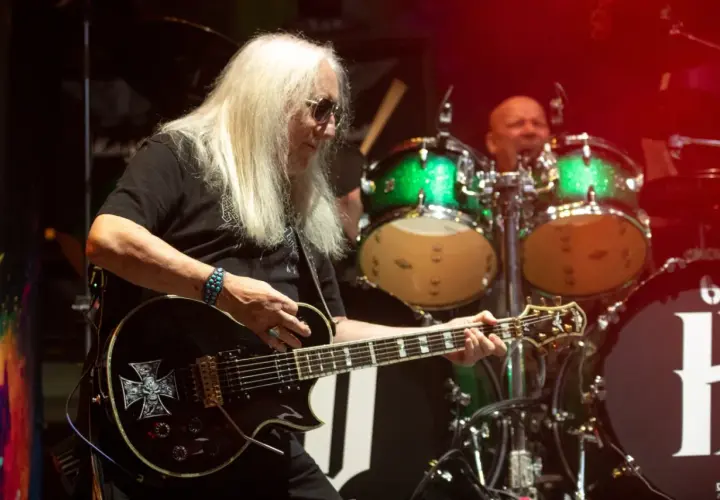 Uriah Heep członkowie na scenie. Gitarzysta z długimi blond włosami gra na czarnej gitarze z symbolem krzyża. W tle perkusista i zielone bębny.