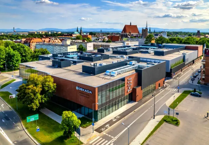 Nowoczesny budynek usługowy "Dekada" z przeszkloną fasadą i ceglanymi elementami, otoczony zielenią i drogami.