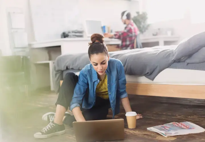 Młoda kobieta pracuje na laptopie na podłodze w pokoju. Obok niej kubek kawy i gazeta. W tle mężczyzna w słuchawkach. Zastanawia się, ile osób może mieszkać w wynajmowanym mieszkaniu.