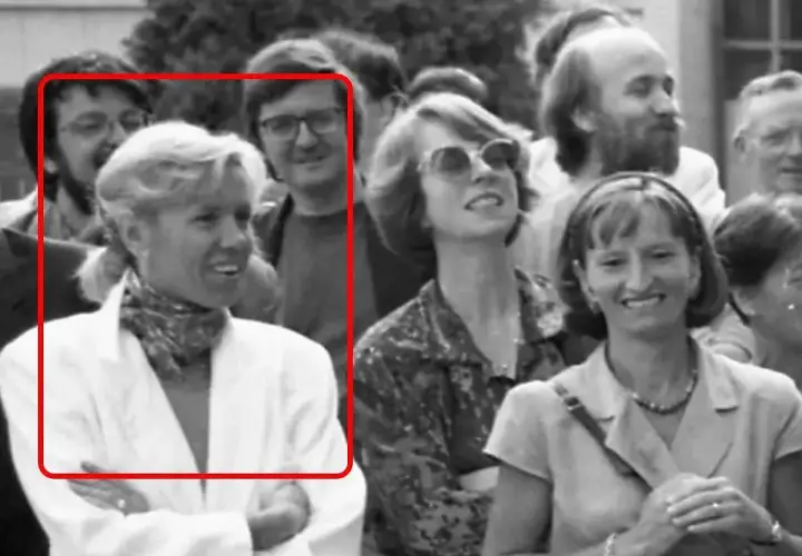 Brigitte Macron młoda, w białej marynarce i z apaszką na szyi, uśmiecha się do aparatu. W tle widać innych ludzi.