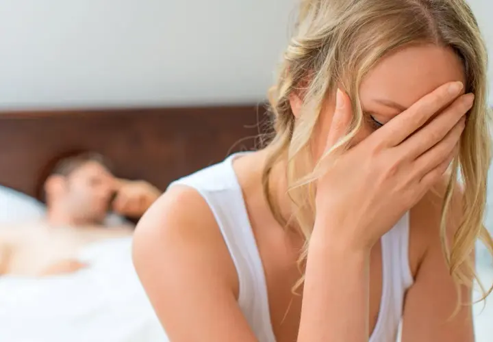 Kobieta zakrywa twarz dłonią, zmartwiona. W tle leży mężczyzna. Zastanawia się, dlaczego nie mam orgazmu.