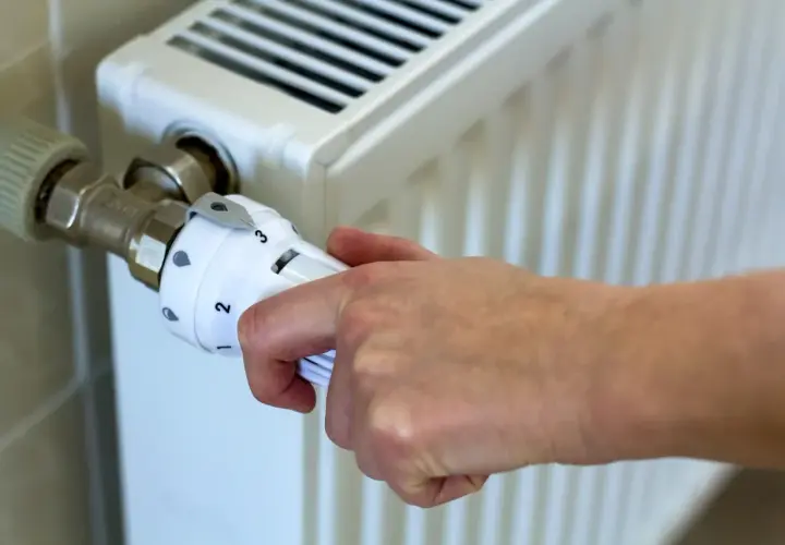 Dłoń reguluje termostat grzejnika, ustawiając temperaturę zgodnie z przepisami dotyczącymi minimalnej temperatury w mieszkaniu.