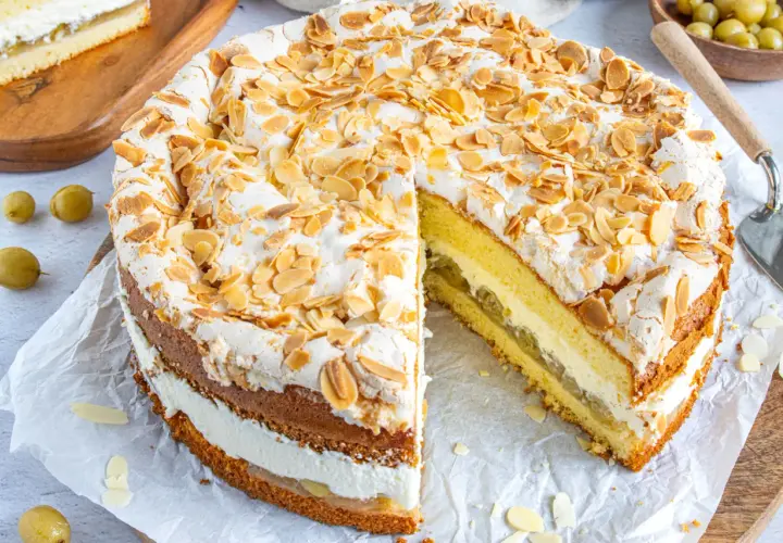 Ein köstlicher Stachelbeer Baiser Kuchen mit Biskuitböden, Sahne und Mandelsplittern. Ein Stück ist bereits angeschnitten.