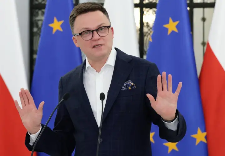 Szymon Hołownia w garniturze przemawia przed flagami Polski i UE.