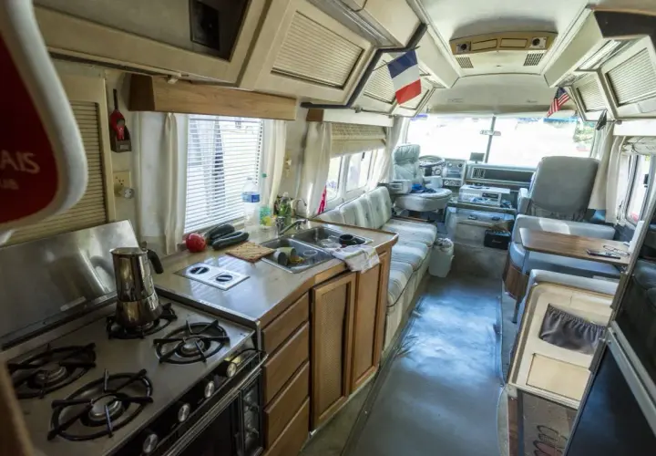 Vue de l'intérieur d'un camping-car américain : cuisine avec cuisinière, évier, et banquette. Drapeaux français et américain visibles.
