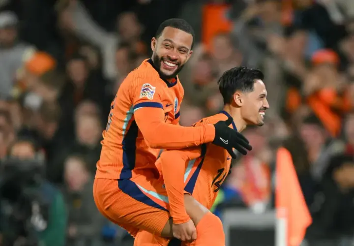 Dwóch piłkarzy Holandii w pomarańczowych strojach świętuje bramkę. Jeden z nich, Memphis Depay, niesie kolegę na plecach, obaj uśmiechnięci.