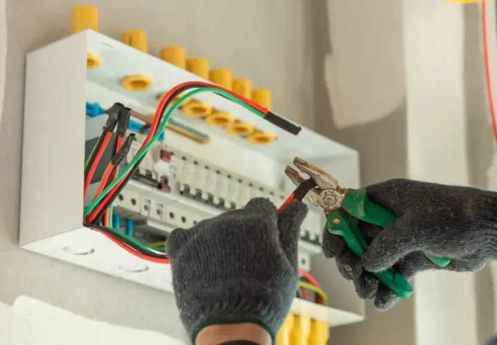 Elektryk w rękawicach pracuje przy skrzynce z bezpiecznikami. Zastanawiasz się, ile zarabia pomocnik elektryka na godzinę?