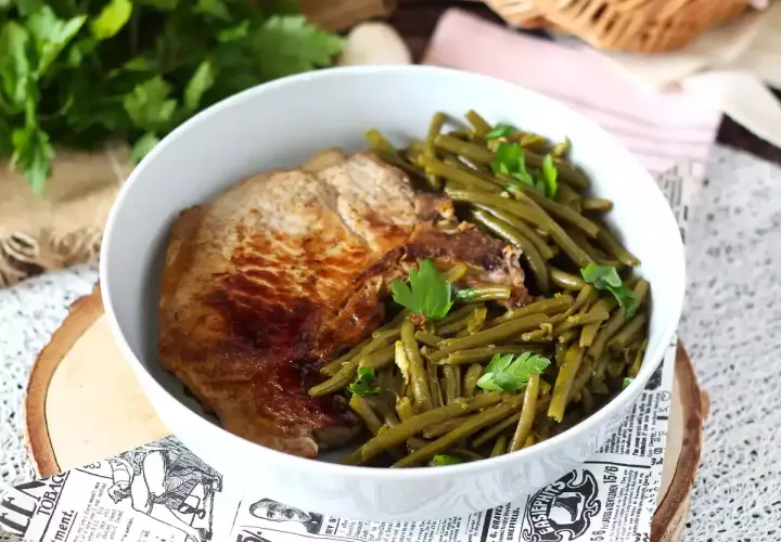 Une savoureuse côte de porc grillée accompagnée de haricots verts frais et persil, servie dans un bol blanc.