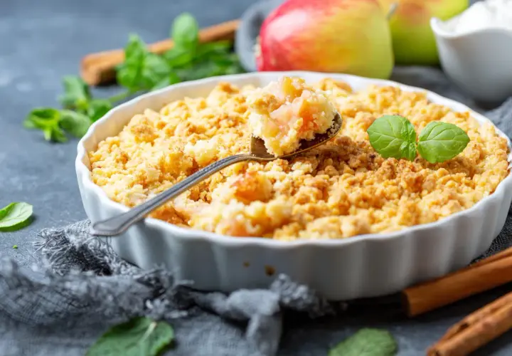 Un délicieux crumble pomme doré, garni de feuilles de menthe fraîche. Des pommes rouges et vertes sont visibles en arrière-plan.