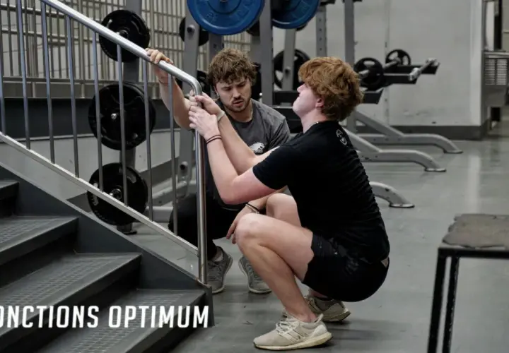 Un coach aide un athlète à améliorer sa **mobilisation articulaire** lors d'un exercice de squat près d'un escalier dans une salle de sport.