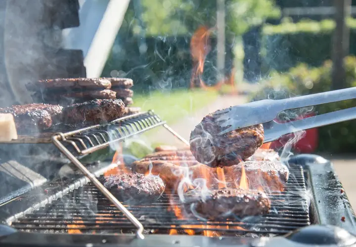 Des hamburgers grillent sur un barbecue à gaz. La fumée s'élève, les flammes dansent. Un hamburger est retourné avec une pince.