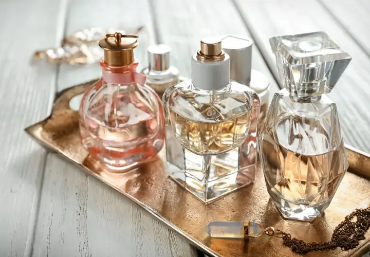 Eleganckie flakoniki perfum, w tym jeden z delikatną różową wstążką, stoją na złotej tacy. W tle błyszcząca biżuteria. Zapach, być może z nutą hexyl cinnamal, unosi się w powietrzu.