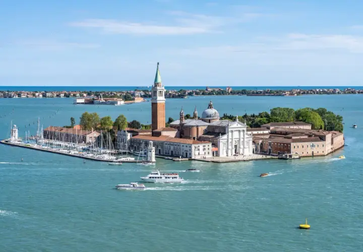 Widok na wyspę San Giorgio Maggiore w Wenecji, z kościołem i dzwonnicą. Wokół pływają łodzie, w tym prom.