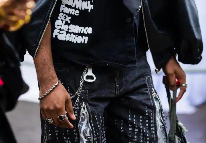 Mężczyzna w czarnej kurtce i spodniach z nadrukiem cyfrowym. Na koszulce widnieje napis "Amour, Fame, Sex, Elegance, Fashion", przypominający opium label.