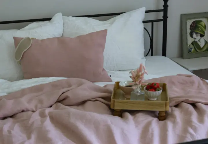 Ein rosafarbenes Leinenbett mit Kissen und einer Augenmaske. Ein Tablett mit Erdbeeren und Kaffee zeigt, wie wird leinen weich.