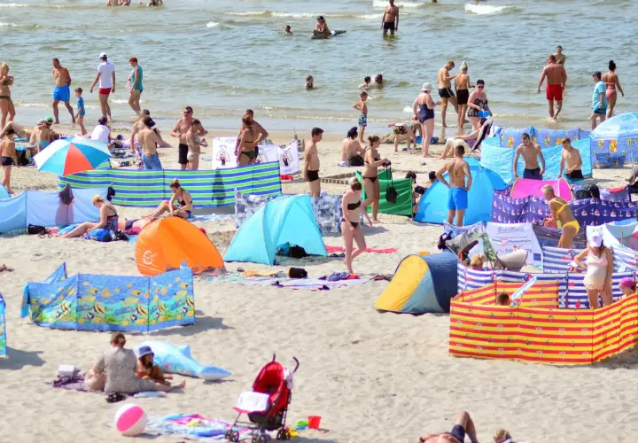 Tłum ludzi na plaży, opalający się i kąpiący w morzu, kolorowe parawany i namioty plażowe.