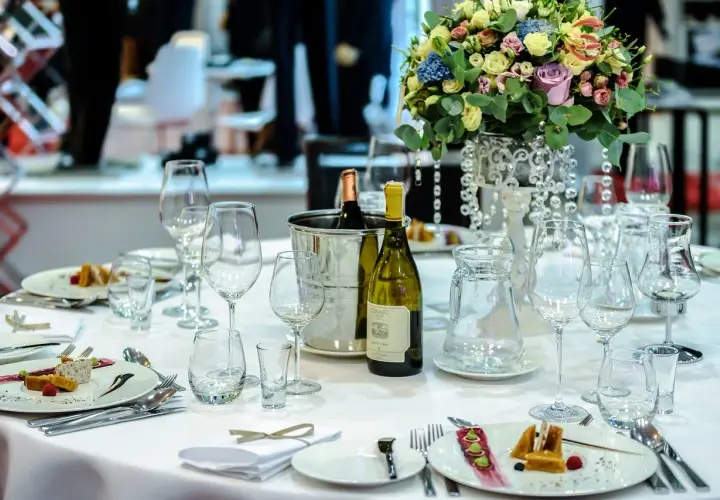 Elegancko nakryty stół w restauracji, gotowy na przyjęcie weselne. Na białym obrusie stoją kieliszki, butelka wina i piękne kwiaty.