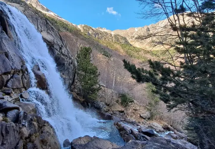 Cascada imponente en un paisaje montañoso, ideal para explorar rutas vielha. El agua cae sobre rocas y vegetación, con árboles y picos nevados al fondo.
