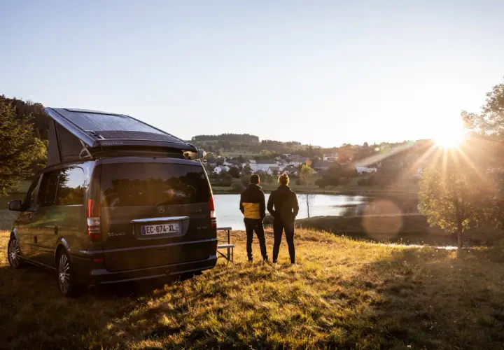 Un camping-car Westfalia avec panneaux solaires, prêt pour l'aventure près de Vulcania. Deux personnes contemplent un lac paisible au coucher du soleil.