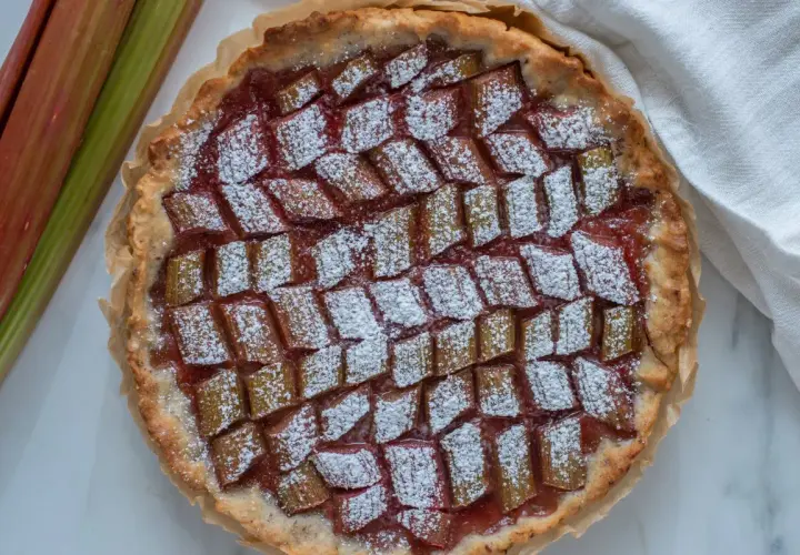 Pyszna tarta z rabarbarem, posypana cukrem pudrem, obok świeże łodygi rabarbaru.
