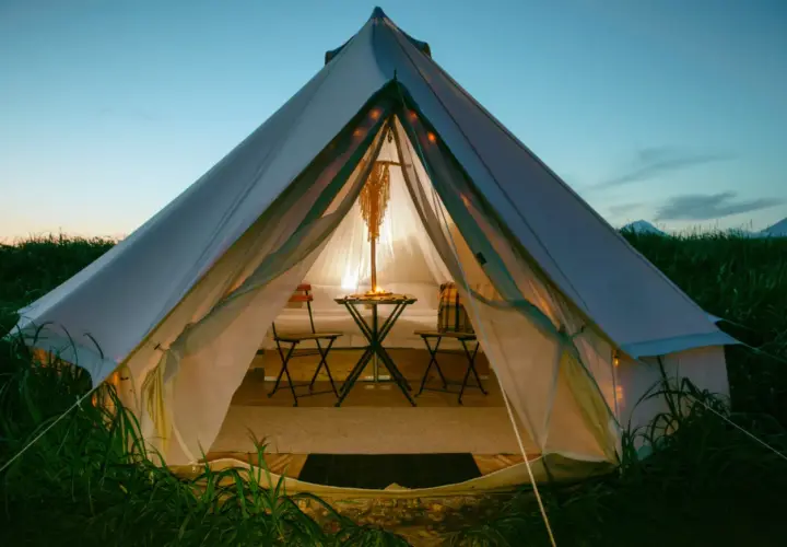 Przytulna glamping jurta oświetlona od środka, z małym stolikiem i krzesłami, otoczona zieloną trawą o zmierzchu.