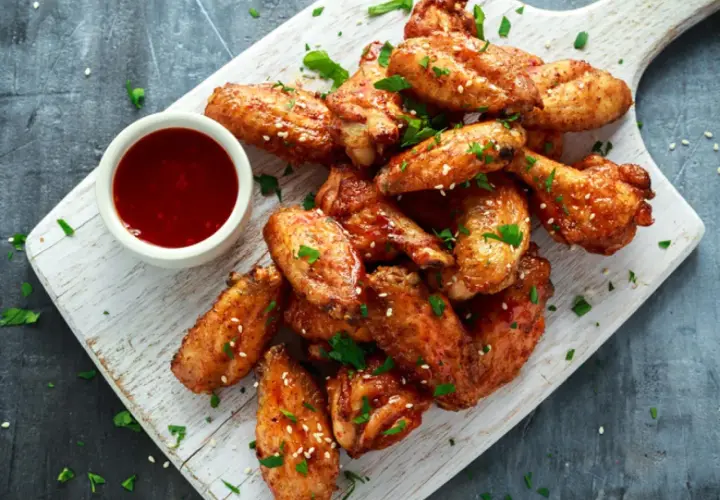 Alitas de pollo al horno jugosas, doradas y espolvoreadas con sésamo y perejil, acompañadas de una salsa roja en un cuenco.