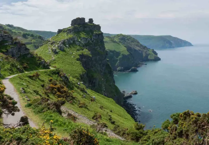 A winding path hugs a grassy cliff overlooking the sea. This is the salt path, a scenic route with rugged beauty.