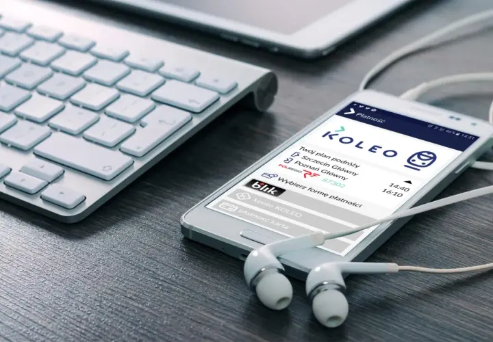 Smartfon z aplikacją KOLEO pokazuje plan podróży i opcje płatności, idealny do wyszukiwania zniżki PKP. Słuchawki i klawiatura sugerują planowanie podróży.