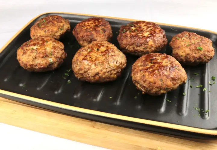 Sieben goldbraune Bouletten, wie nach Omas Rezept, liegen auf einem schwarzen Grillteller, garniert mit Petersilie.